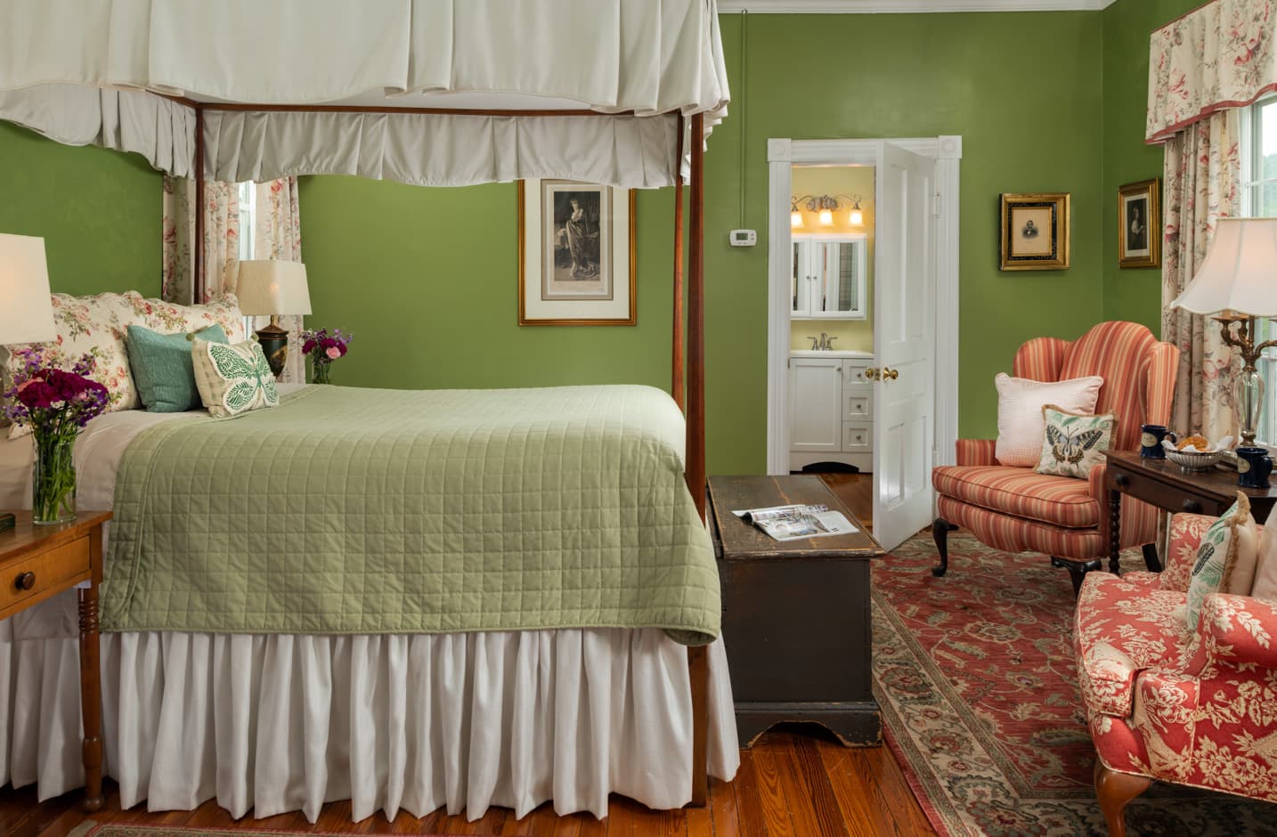 Canopied four-poster queen bed with a green blanket in a room with hardwood floors and a seating area