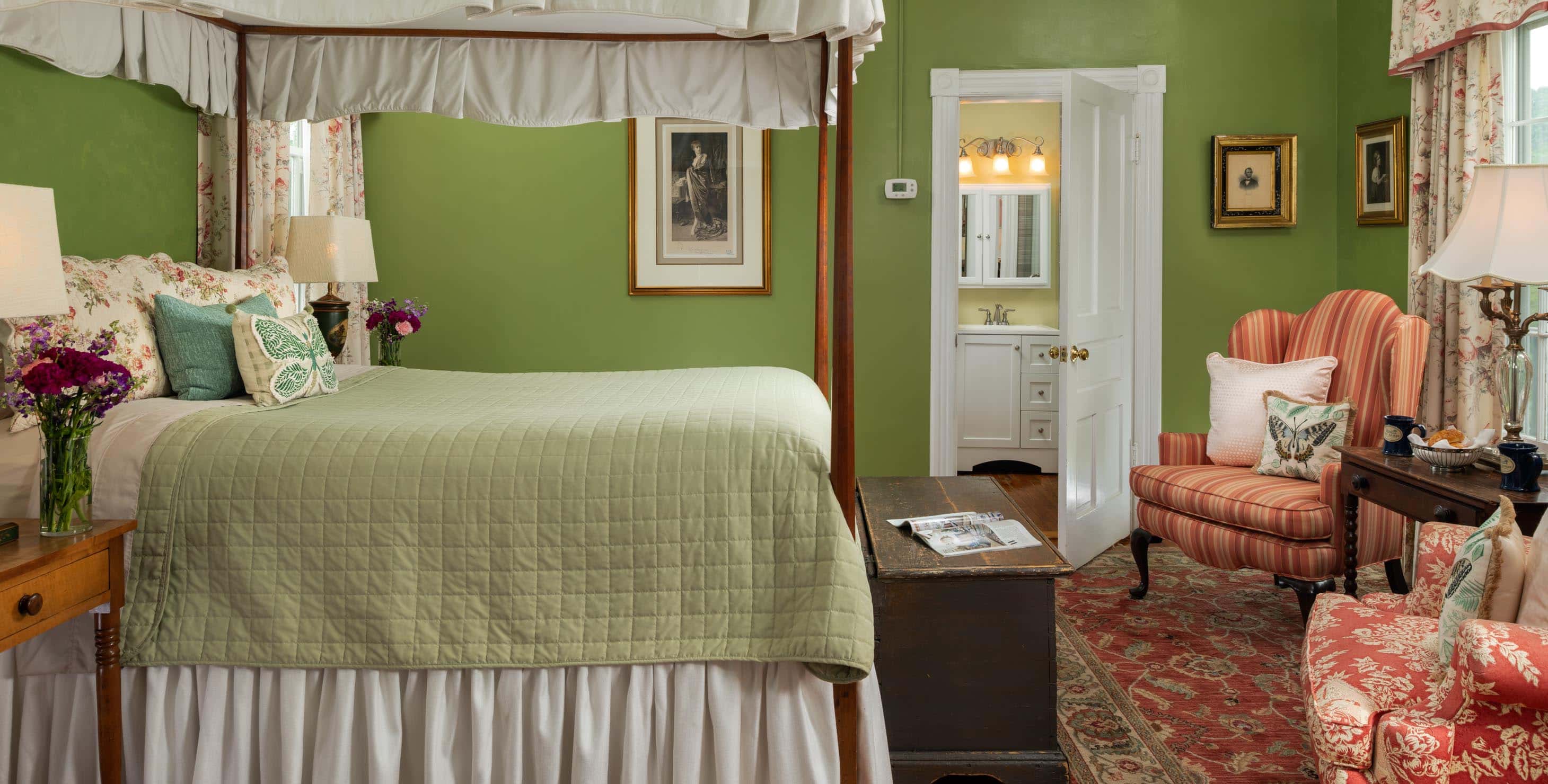 Canopied four-poster queen bed with a green blanket in a room with hardwood floors and a seating area