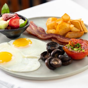 Two eggs sunny side up with bacon, potatoes, mushrooms, and tomato